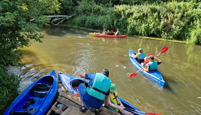 Rodzina w kajakach na rzece, przygotowująca się do spływów kajakowych Prosną. Zielone brzegi i słoneczny dzień.